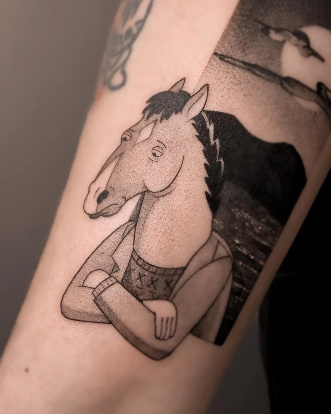 Tatuaż przedstawiający postać BoJacka Horsemana na tle księżycowego pejzażu, wykonany na przedramieniu.