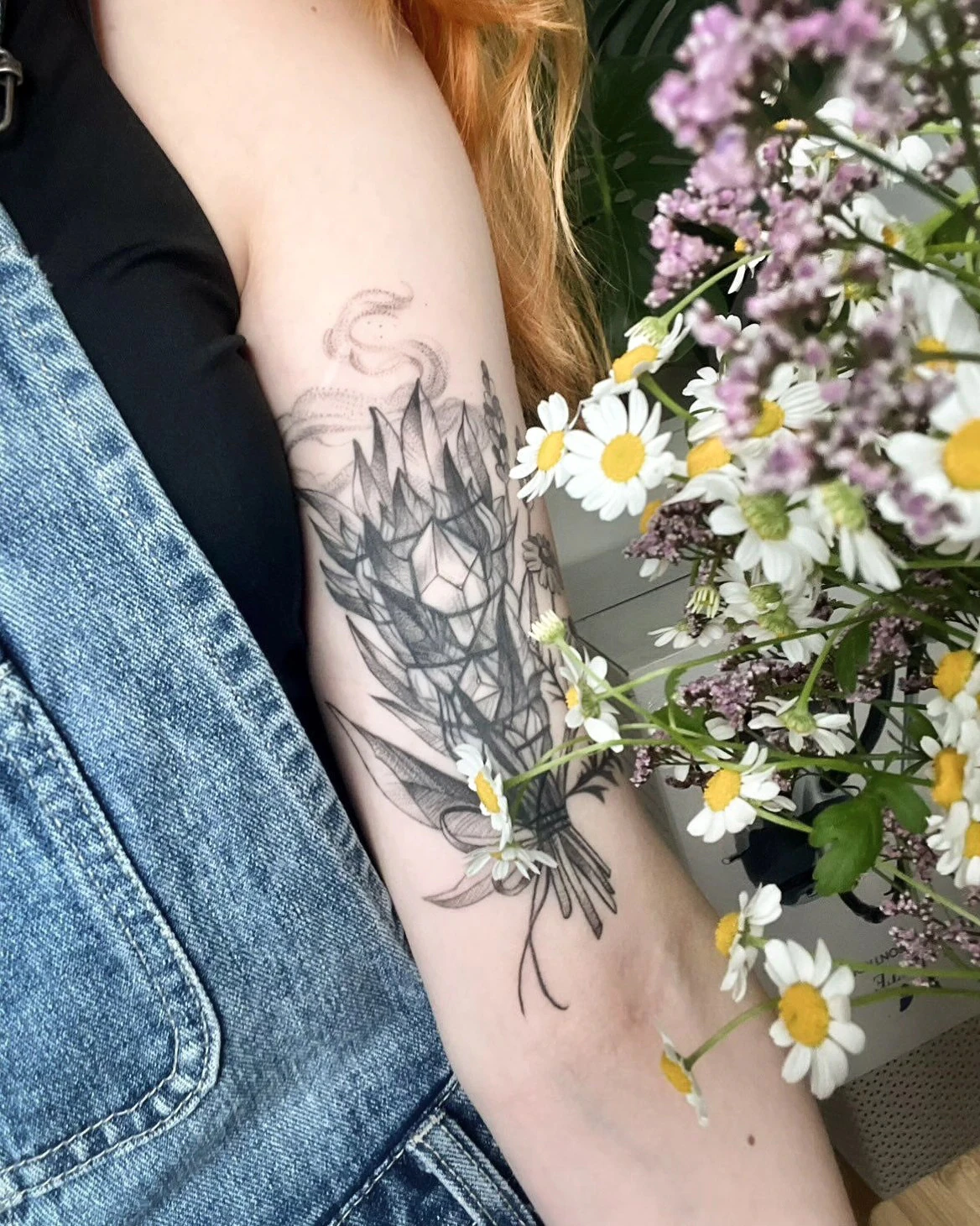 Wygojony cover tatuażu na ramieniu, przedstawiający botaniczną kompozycję z liśćmi, kwiatami i ornamentem; obok widoczny bukiet polnych kwiatów.