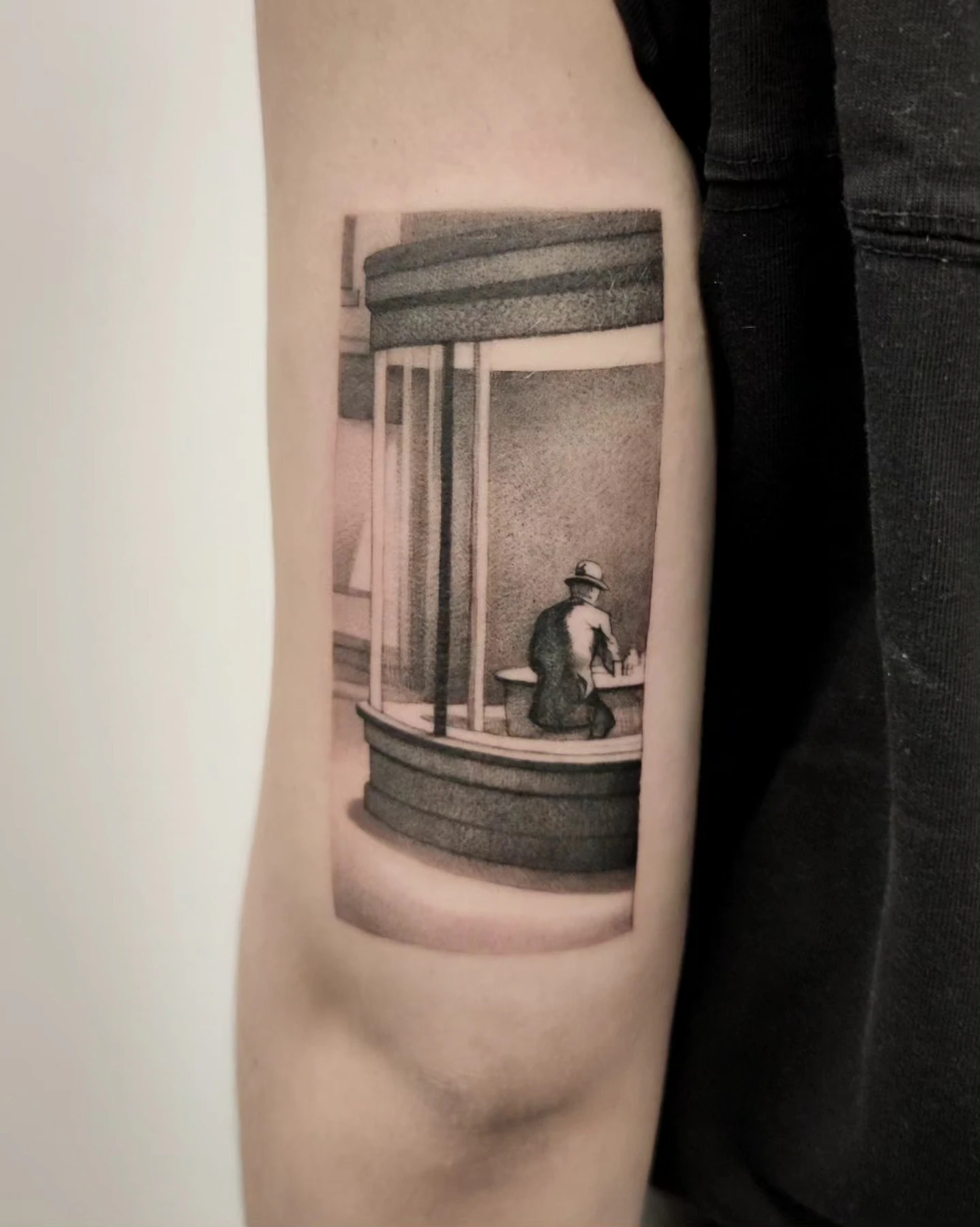 Tatuaż przedstawiający osobę siedzącą przy oknie lub witrynie, wykonany w monochromatycznej tonacji na podstawie obrazu Edwarda Hoppera.