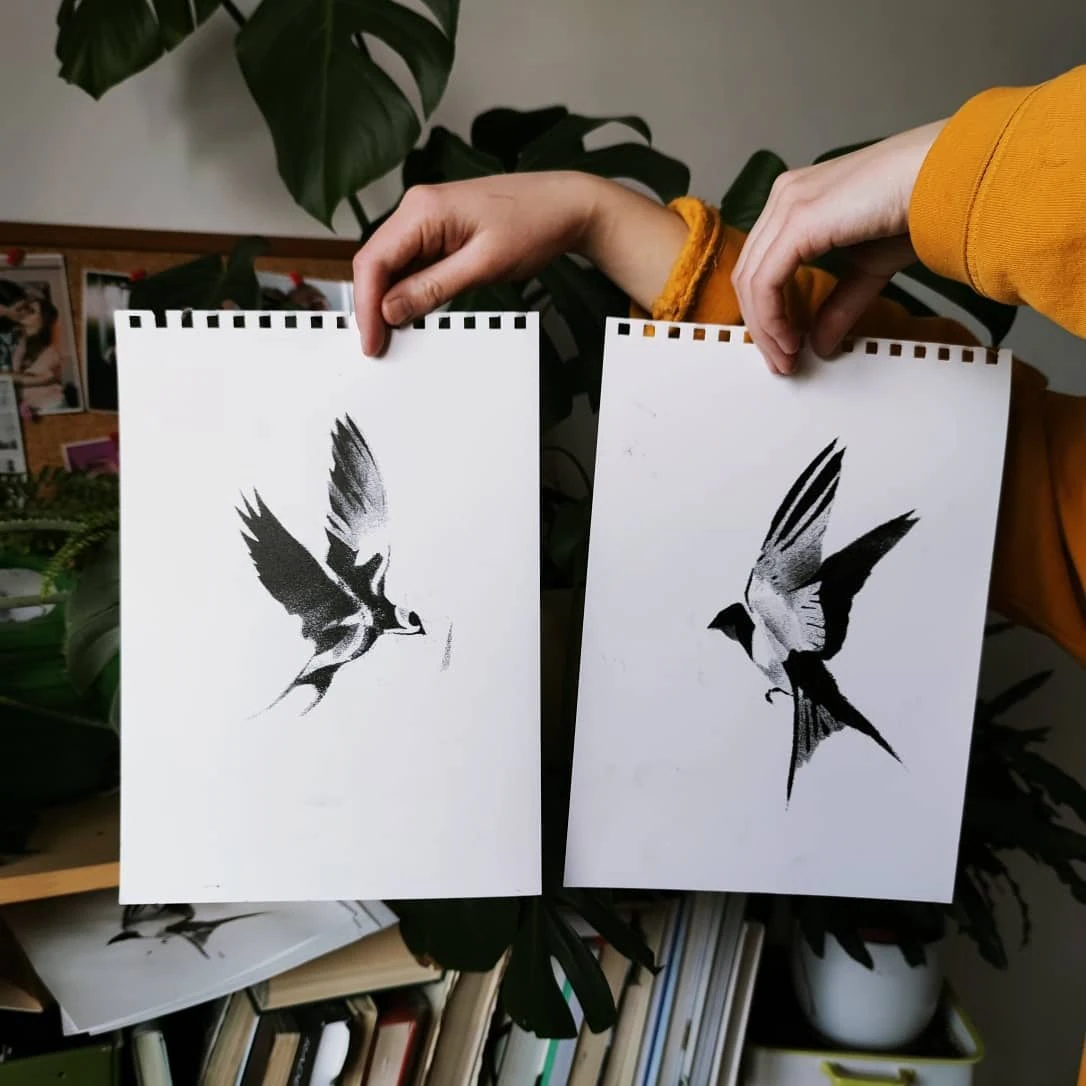 Ręcznie wykonane czarno-białe printy z wizerunkami jaskółek na białym papierze, prezentowane w dłoniach na tle roślin i pracowni.
