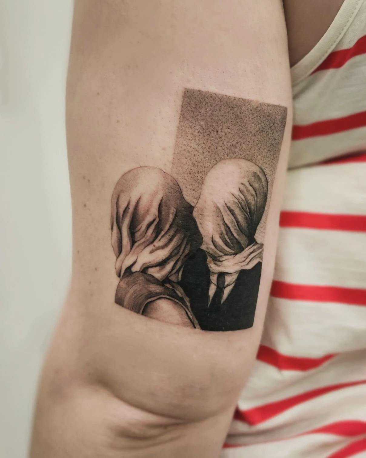 Zbliżenia tatuażu przedstawiającego dwie postacie z zasłoniętymi twarzami, inspirowanego obrazem „Kochankowie” René Magritte’a.