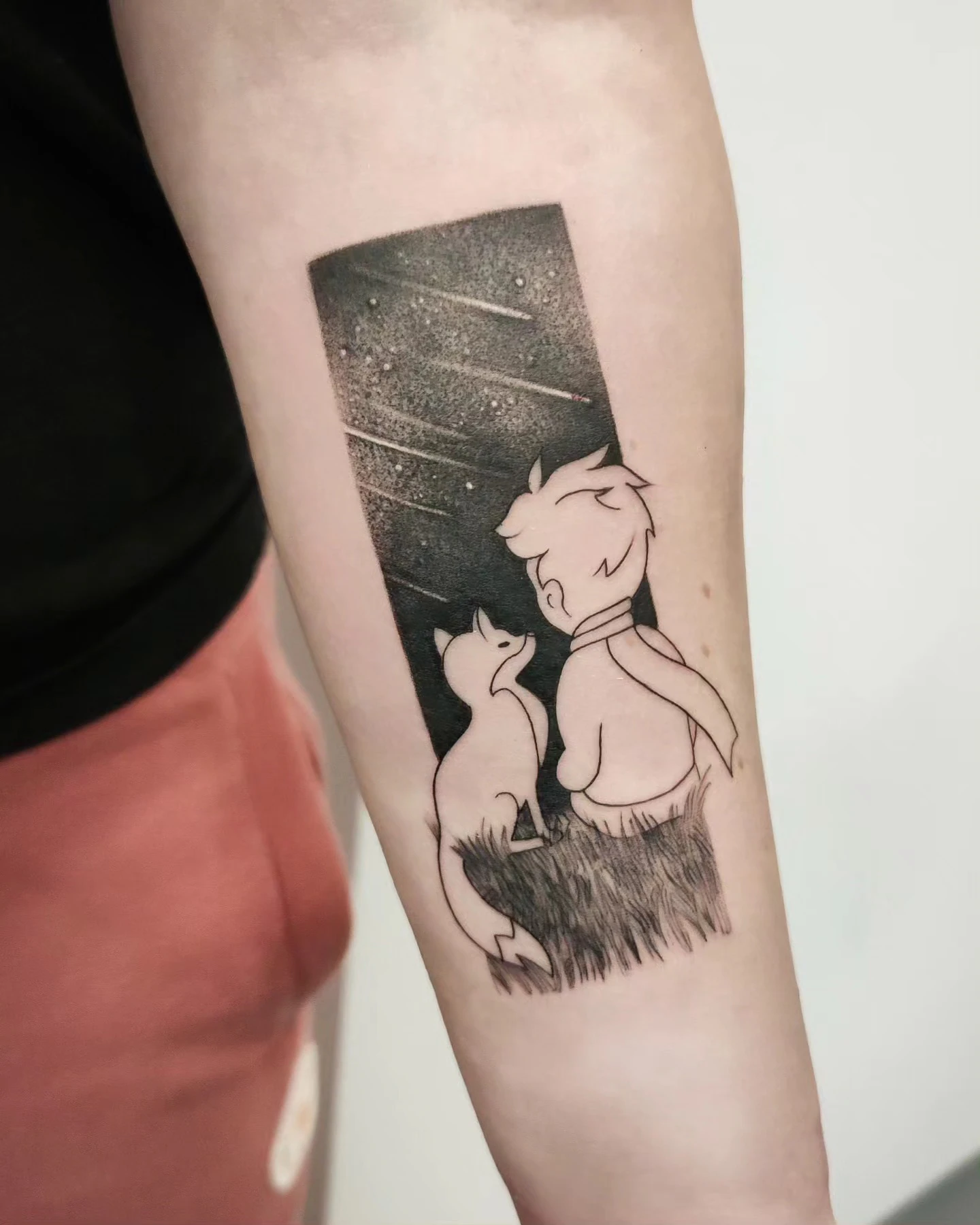 Tatuaż przedstawiający Małego Księcia i lisa siedzących na trawie pod gwiaździstym niebem.