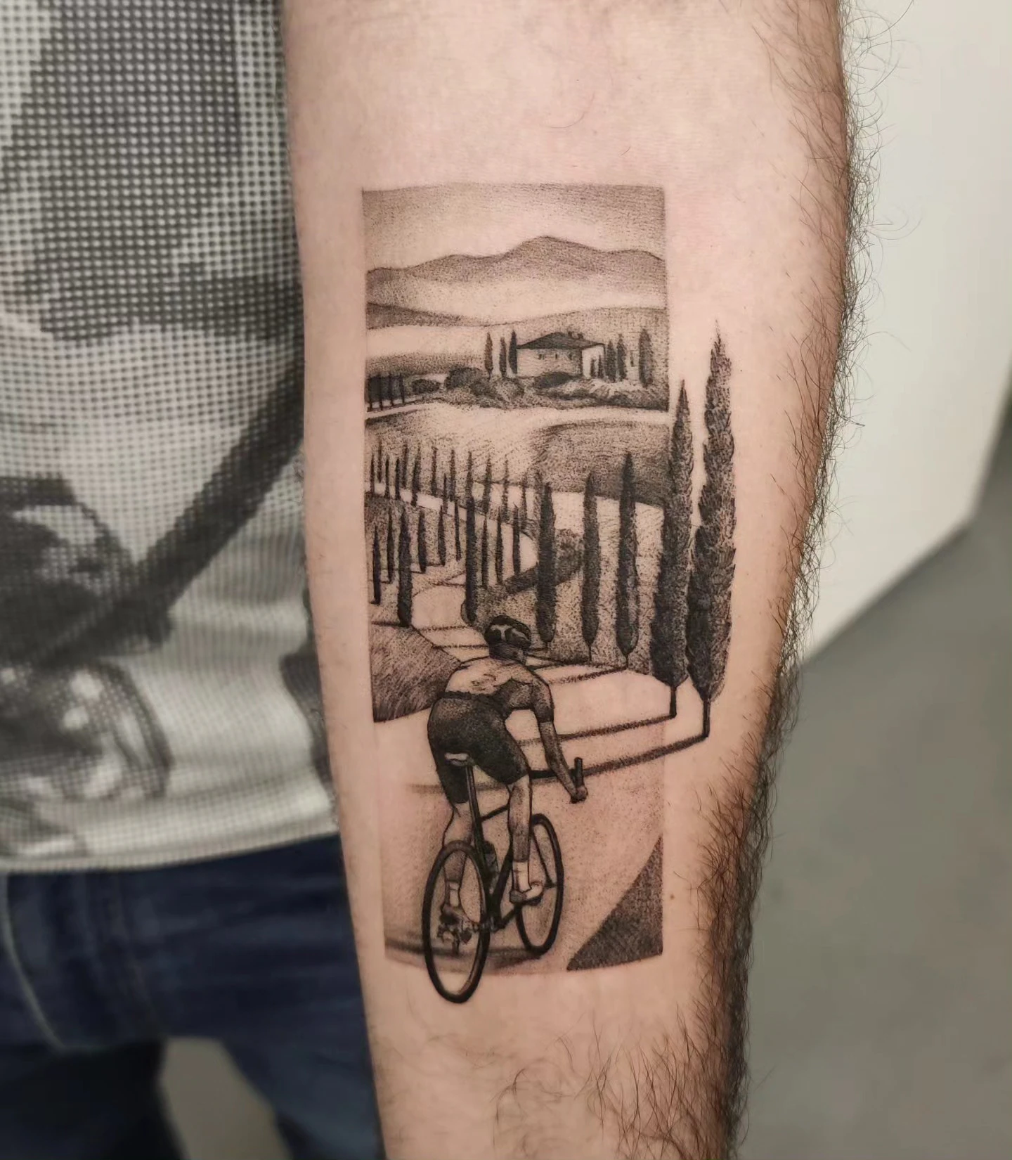 Tatuaż przedstawiający rowerzystę jadącego przez krajobraz z cyprysami, wzgórzami i drogą, wykonany na przedramieniu.