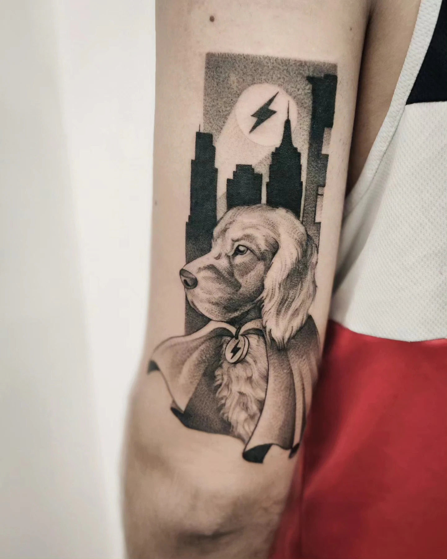 Tatuaż przedstawiający spaniela w stroju superbohatera, z emblematem błyskawicy i miejską panoramą w tle.