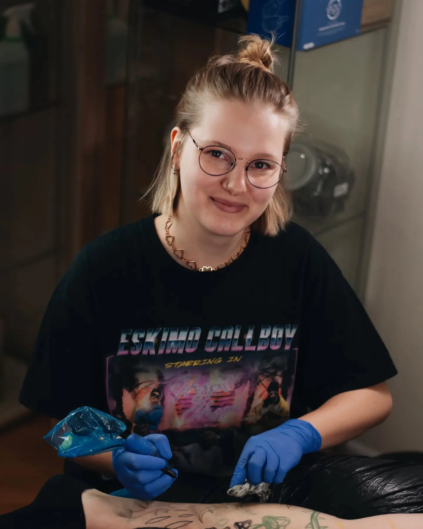 Klara Keczyńska w okularach i niebieskich rękawiczkach pracuje nad tatuażem; na kolejnych ujęciach pozuje z maszynką do tatuowania.