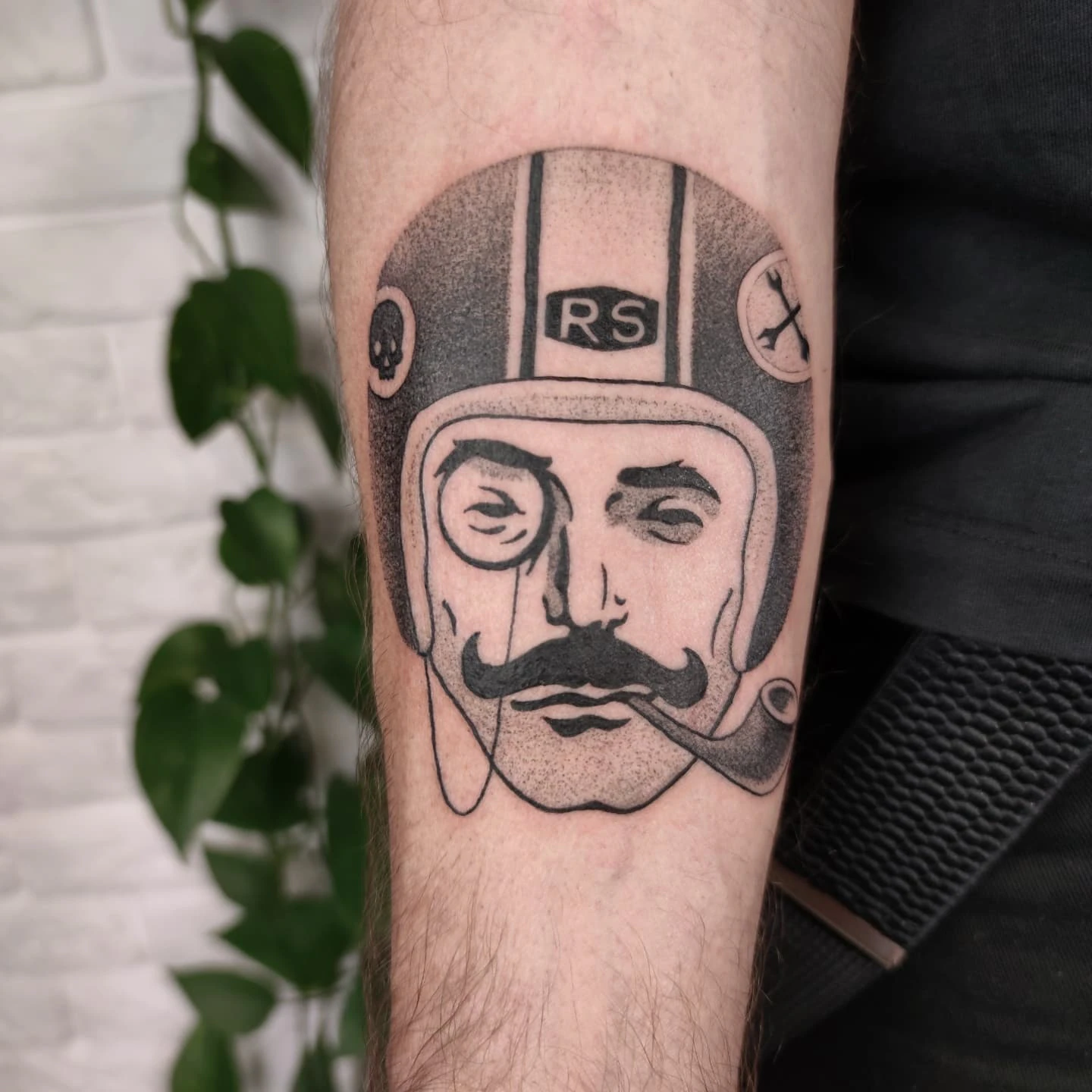 Tatuaż na przedramieniu przedstawiający stylizowaną twarz w kasku motocyklowym, z monogramem „RS” powyżej; obok mężczyzna prezentuje tatuaż siedząc na motocyklu.