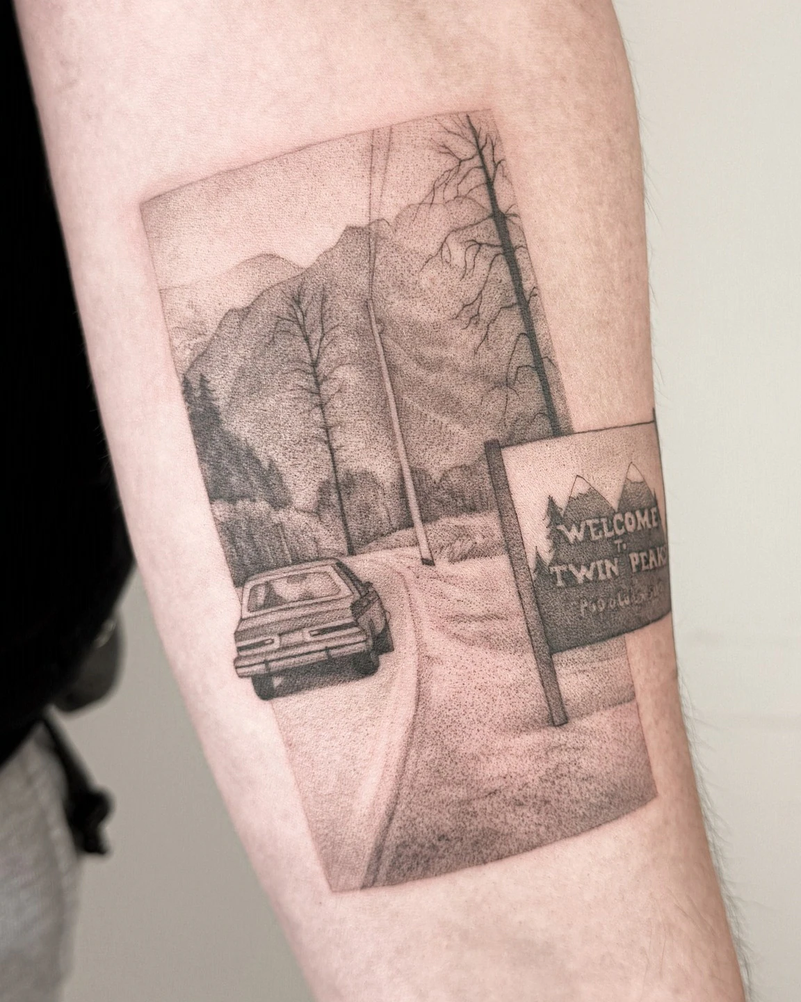 Tatuaż przedstawiający drogę prowadzącą przez górski krajobraz, samochód oraz znak „Welcome to Twin Peaks” na ramieniu.
