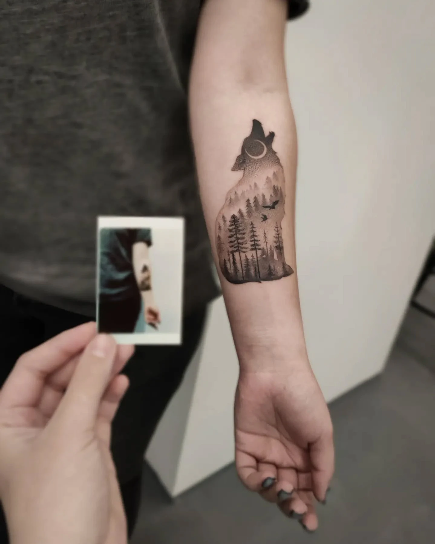 Tatuaż leśnego wilka na przedramieniu, z sylwetką wilka, księżycem, drzewami i ptakami; na części zdjęć widoczny wydruk fotografii tego tatuażu.